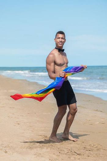 Man holding a rainbow gay pride flag while standing on the beach.