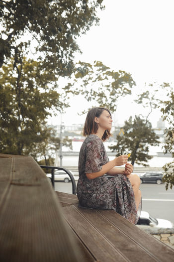 Young brunette woman in long dress sits on city wooden bench and holds an autumn leaf in her hand