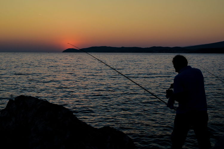 Rear view of man fishing in sea against sunset sky