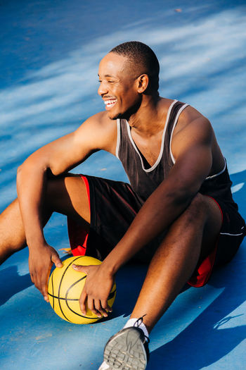 Full body of young african american athlete in sportswear sitting on floor of sports ground with yellow basketball in daylight