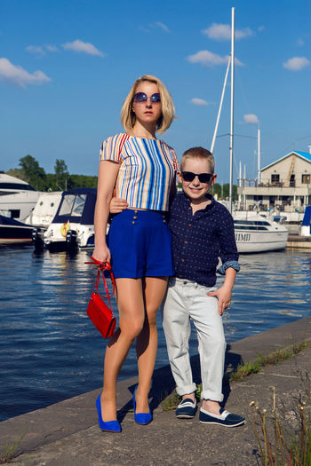 Young business woman in short blue trunks standing at the pier with yachts and boats and her son