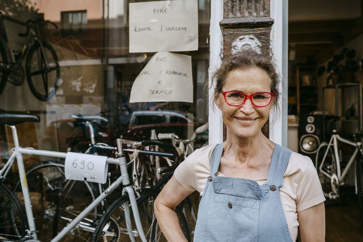Portrait of confident female owner wearing funky eyeglasses while standing against bicycle shop