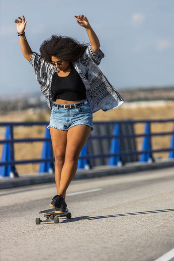 Young and afro woman skating long board by an empty bridge at sunset, front view