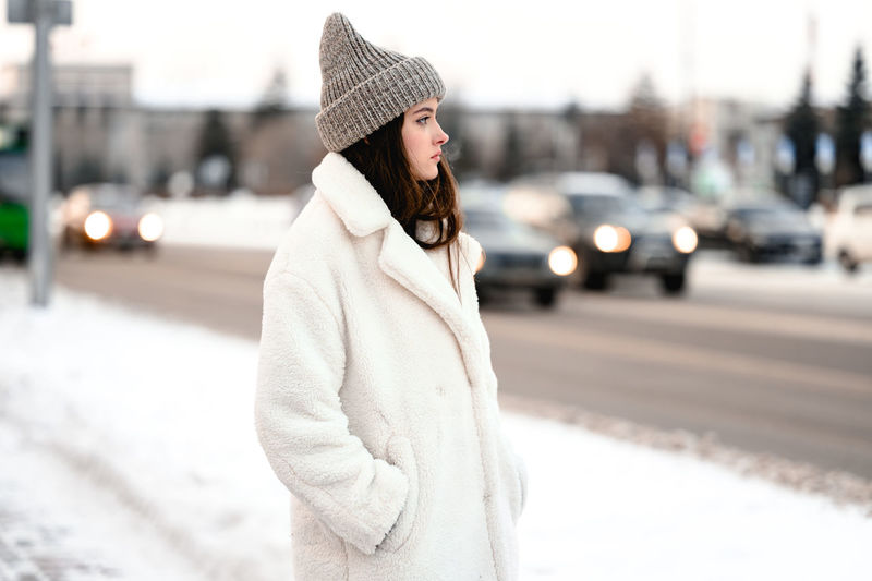 Girl in a fur coat walks along the road traffic of cars. the girl has a knitted hat on her head