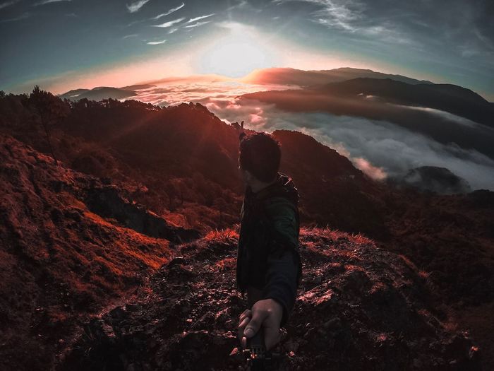 Fish-eye lens view of man standing on mountains against sky during sunset