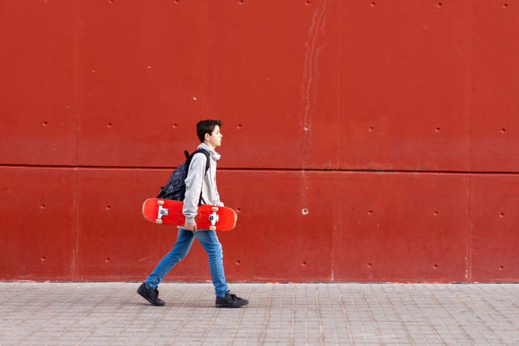 Young boy holding a red skateboard while walking against red wall person