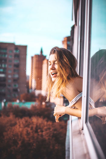 Side view of young woman leaning on window against buildings in city