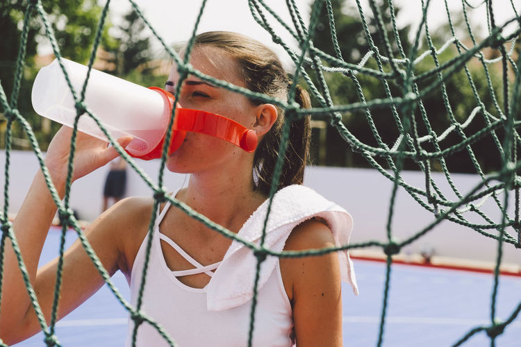Young woman drinking water from bottle on soccer court seen through net