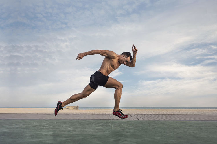 Side view of shirtless man taking running stance against sky