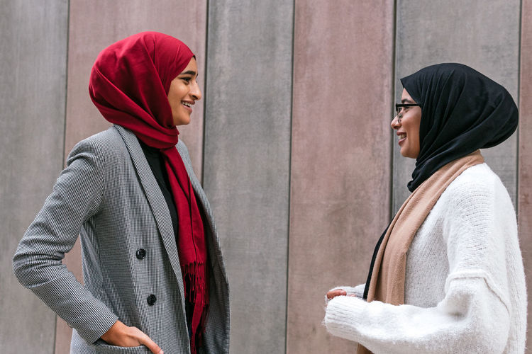 Side view of arab female friends in traditional hijab standing together talking on the street and looking at each other