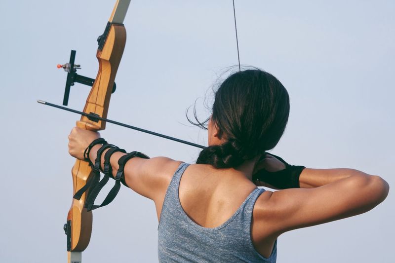 Rear view of woman aiming bow against sky