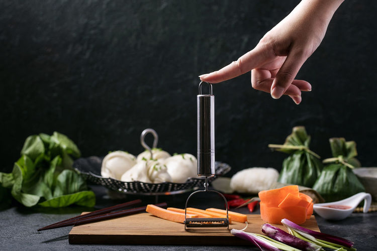 Cropped hand with peeler by vegetables on table against wall