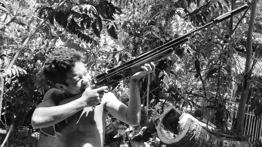 Young man aiming rifle while standing against plants