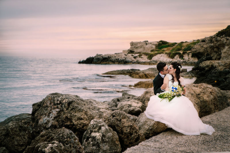 Wedding couple kissing while sitting at rocky beach
