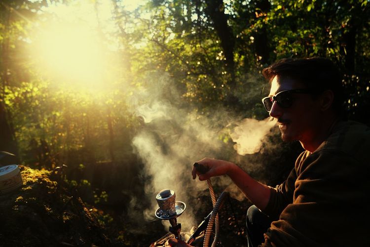 Side view of young man smoking hookah in forest during sunset