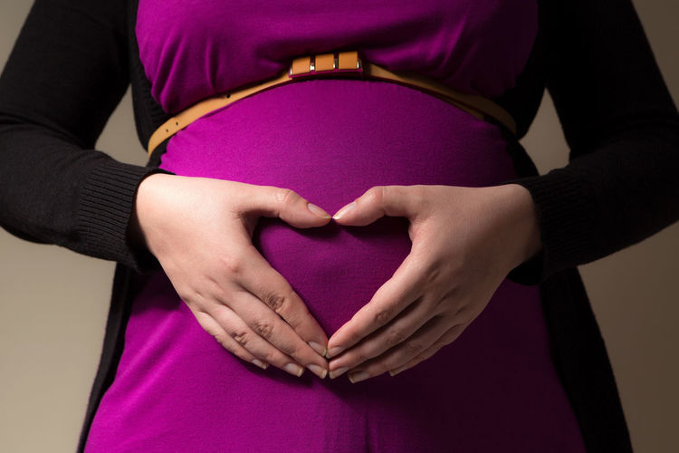 Midsection of pregnant woman making heart shape with hands on stomach against wall
