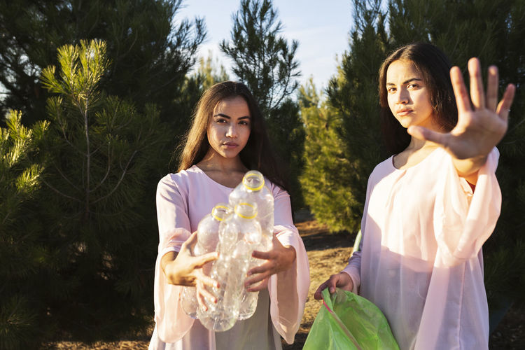 Angry woman making stop gesture standing by sister with empty plastic bottles in forest