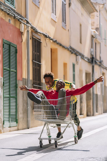 Happy carefree young african american friends in casual clothes riding around in shopping trolley in street