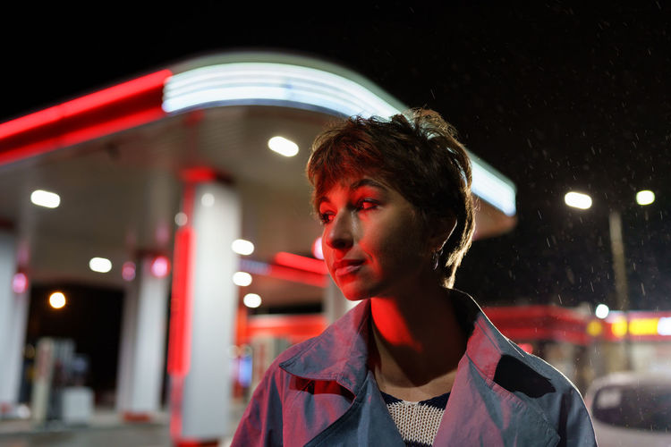 Caucasian woman with red neon light on face posing standing on night street in rainy weather