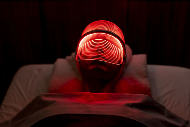 Young woman wearing illuminated red mask during rejuvenating treatment in dark at beauty spa