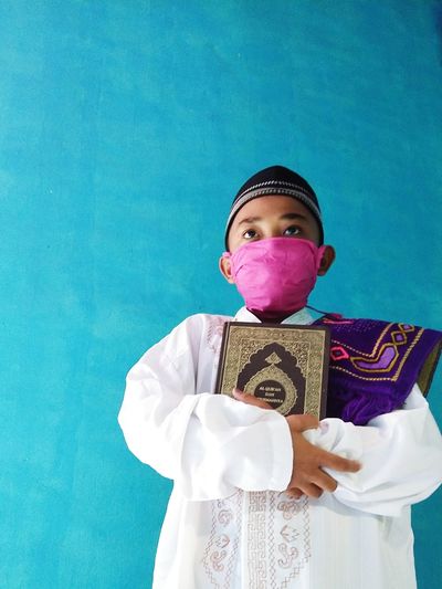 Low angle view of boy wearing mask holding koran while standing against wall