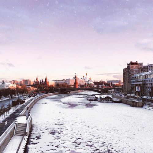 Frozen river in city against sky during sunrise