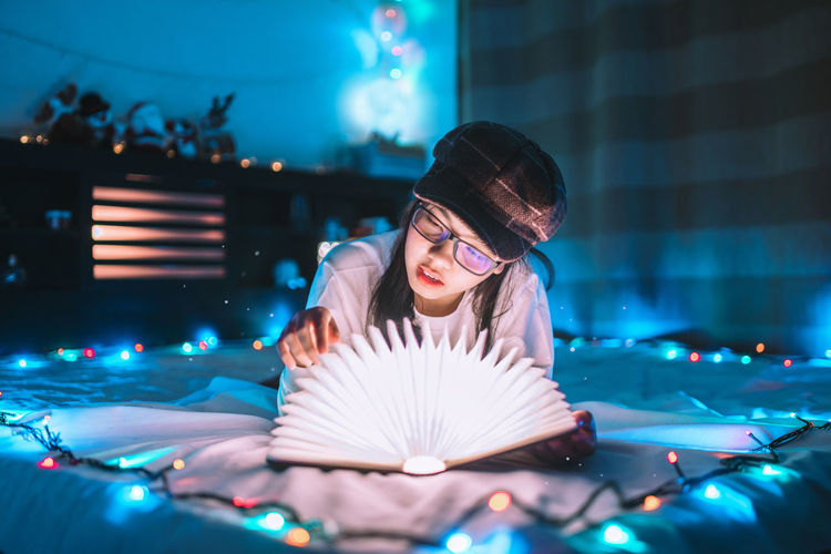 Beautiful young woman reading book amidst lights on bed at night