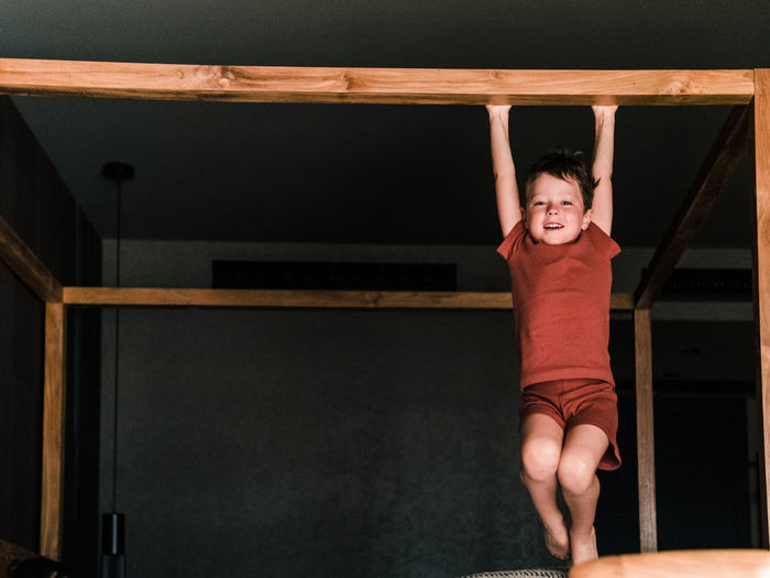 Positive little kid hanging on wooden canopy bed while having fun in bedroom and looking at camera