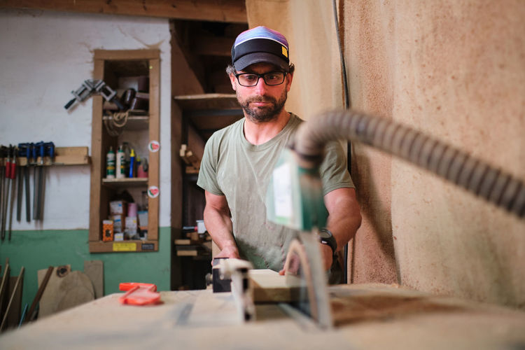 Concentrated craftsman in eyewear cutting wooden plank on table saw against assorted tools in workshop