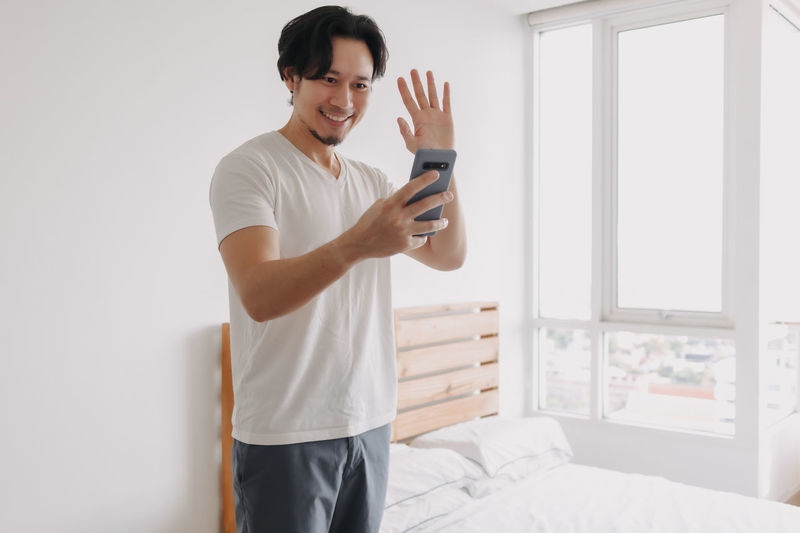 Young man using mobile phone while standing on bed