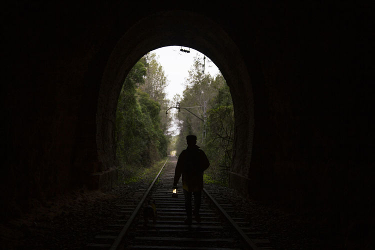 Silhouette of anonymous tourist with lamp walking on rails and admiring forest in dark tunnel in countryside