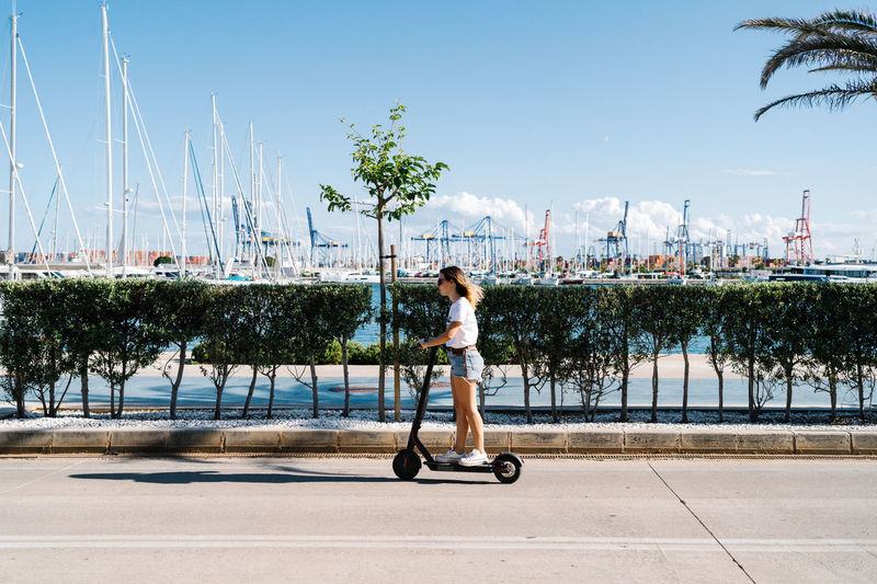 Full body side view of young female in casual outfit riding electric scooter on paved road near urban embankment in sunny summer day