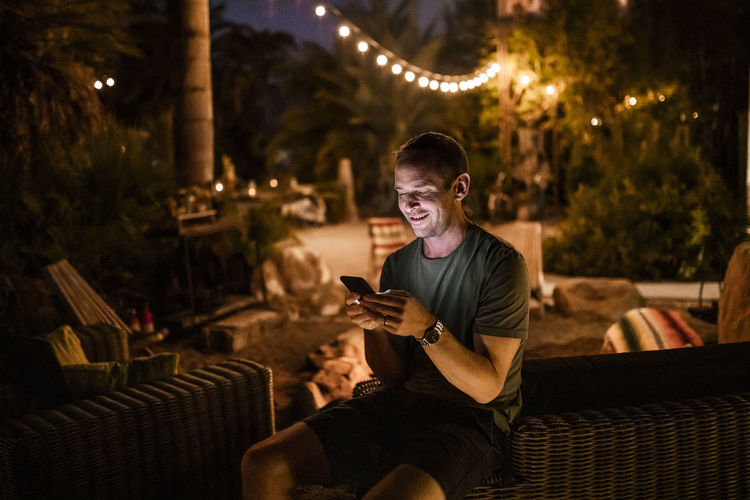 Smiling man using smart phone while sitting in garden during sunset