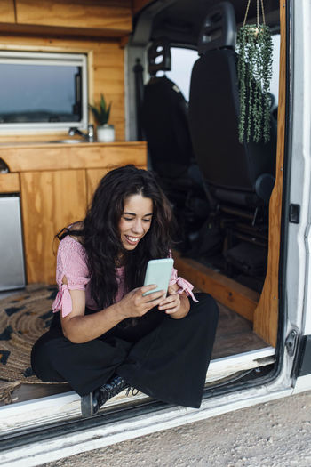 Happy young woman using smart phone in van parked on dirt road