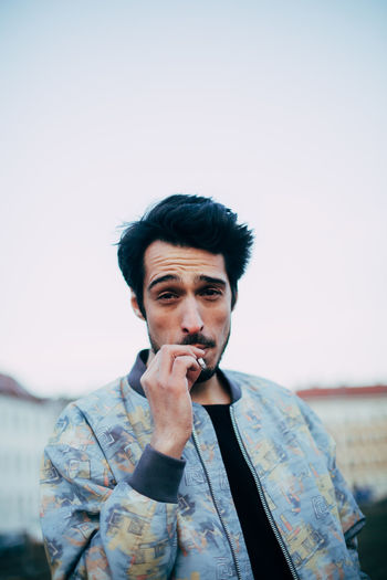 portrait of cool latino man smoking and looking in camera Cool Smoke Smoking Stylish Adults Only Bad Habit Casual Clothing Cigarette Coolness Day Focus On Foreground Front View Headshot Leisure Activity Lifestyles Looking At Camera One Person Outdoors People Portrait Real People Sky Standing Style Young Adult