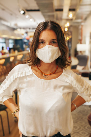 Serious young female employee in casual outfit and protective mask looking at camera while standing against blurred interior of modern coworking space