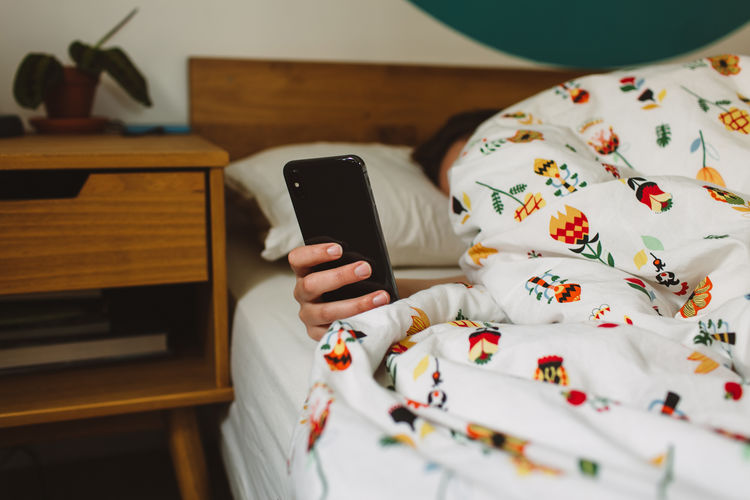 Unrecognizable female checking time on smartphone while lying under blanket in cozy bed in morning