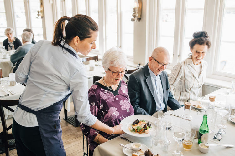 Owner serving food to senior friends sitting by restaurant table
