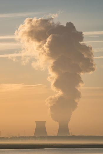 Smoke emitting from nuclear power station against cloudy sky during sunset