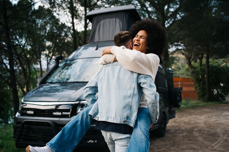 Cheerful young african american woman laughing happily and embracing boyfriend while having fun together near camper van parked in green forest during summer journey together
