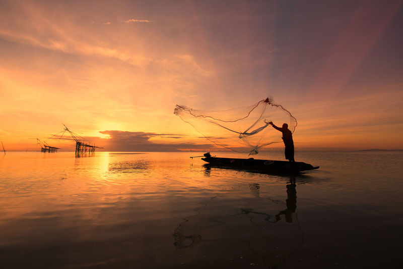 Fisherman with fishing net on boat in sea during sunset 