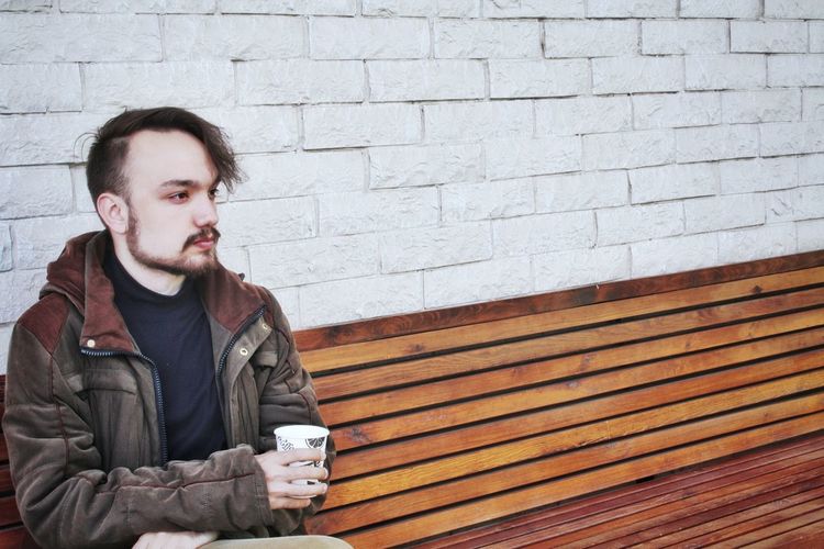 Thoughtful young man holding coffee cup while sitting on bench