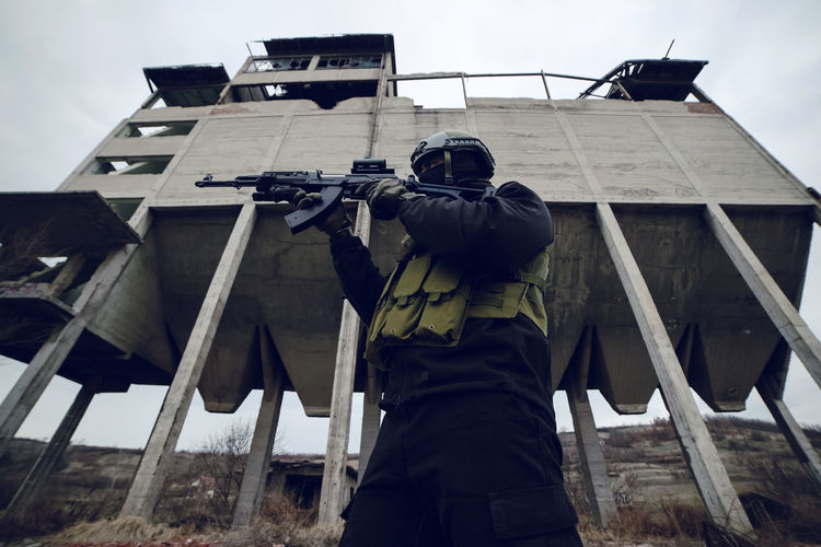 Low angle view of male soldier aiming gun while standing against built structure