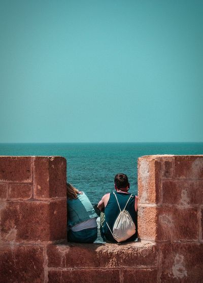 View of people sitting on wall by sea against clear sky