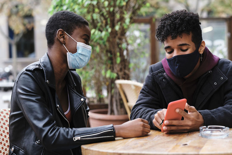 Friends wearing protective face mask using mobile phone while sitting at sidewalk cafe