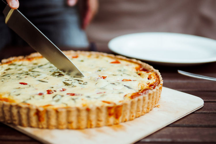 Cropped hand cutting quiche with tomatoes on cutting board on table