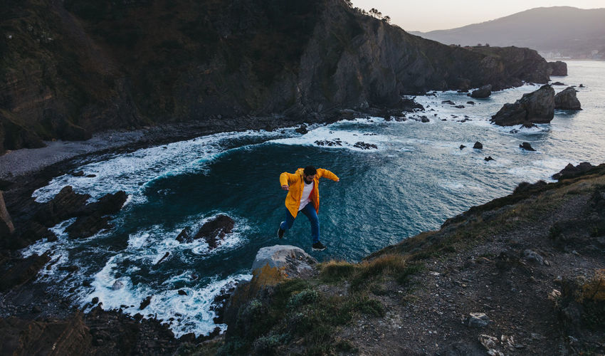 From above person in vibrant yellow jacket jumping on edge of cliff and enjoying amazing scenery of rocky sea coast during sunset in spain