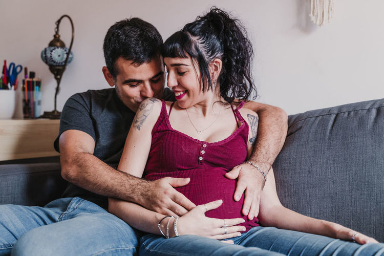 Male kissing and embracing pregnant woman while sitting on sofa at home