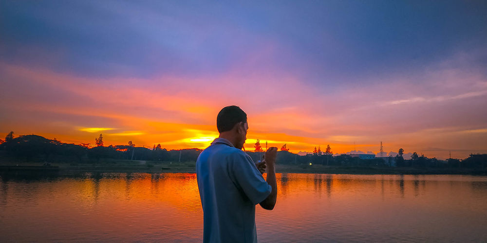Rear view of man standing by lake against sky during sunset