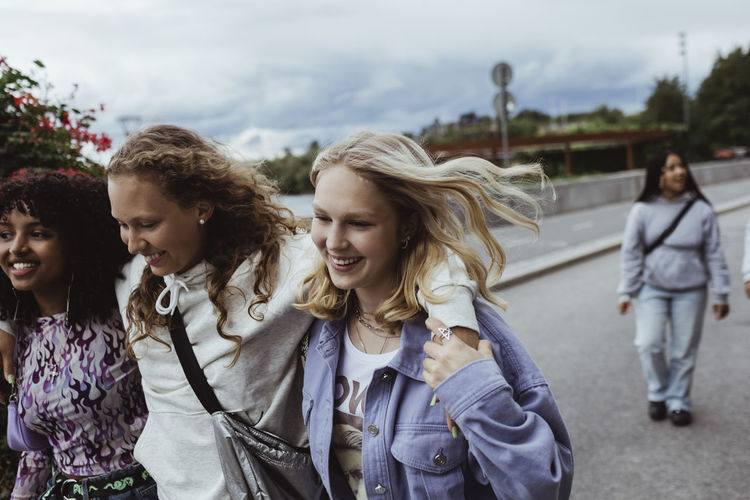Smiling friends with arms around walking on footpath against sky